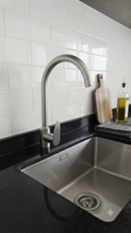 A modern stainless steel high-arc kitchen faucet installed in a black countertop sink with a white subway tile backsplash.